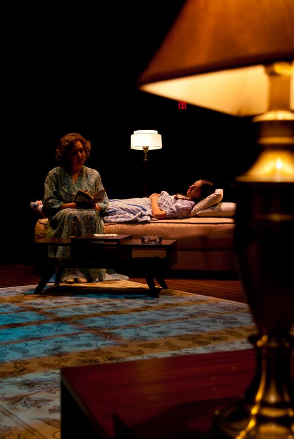 A dimly lit room with a woman sitting on a chair reading a book. Another person lies on a bed under a blanket. A lamp in the foreground casts a warm glow, illuminating the patterned rug beneath.