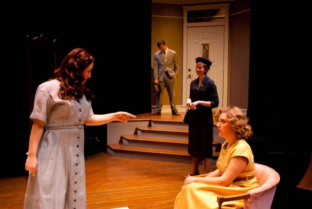 Four people are on a stage set resembling a living room. A woman in a gray dress points as she stands. A woman in a yellow dress sits in a chair. Another woman in black and a man in a suit stand in the background near stairs.