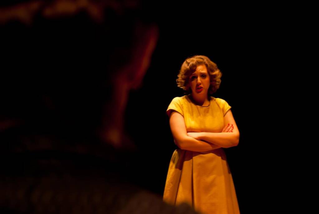 A woman in a yellow dress stands on a dimly lit stage, arms crossed, looking toward someone in the foreground. The background is dark, highlighting her presence.