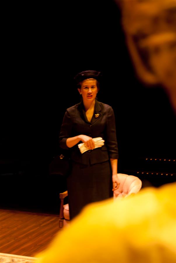 A person in a dark suit and hat stands on stage holding white gloves. They appear to be performing in a theatrical scene, with a blurred foreground figure partially visible. A pink chair is in the background amidst the dark setting.