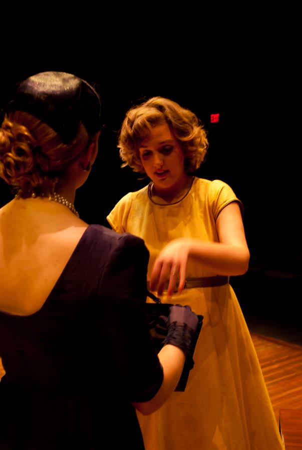 Two women in a dimly lit setting. One is wearing a yellow dress and gesturing with her hands. The other, facing away, is in a dark dress and hat. The scene suggests a theatrical performance or a dramatic interaction.