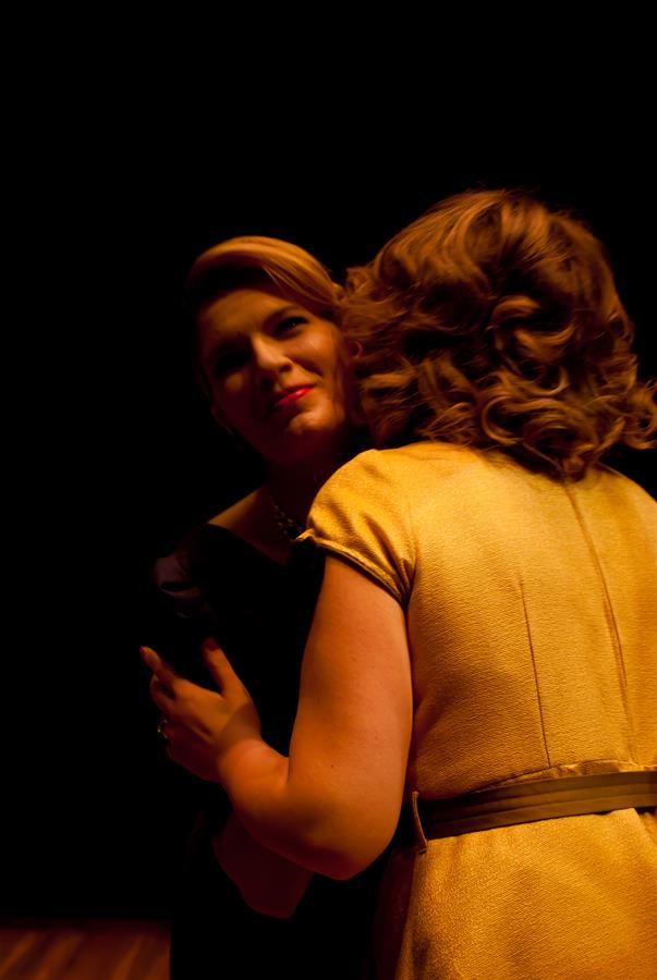 Two women embrace against a dark background. The woman on the left is visible, wearing dark clothing and looking slightly towards the camera. The woman on the right has her back turned, dressed in a yellow outfit.