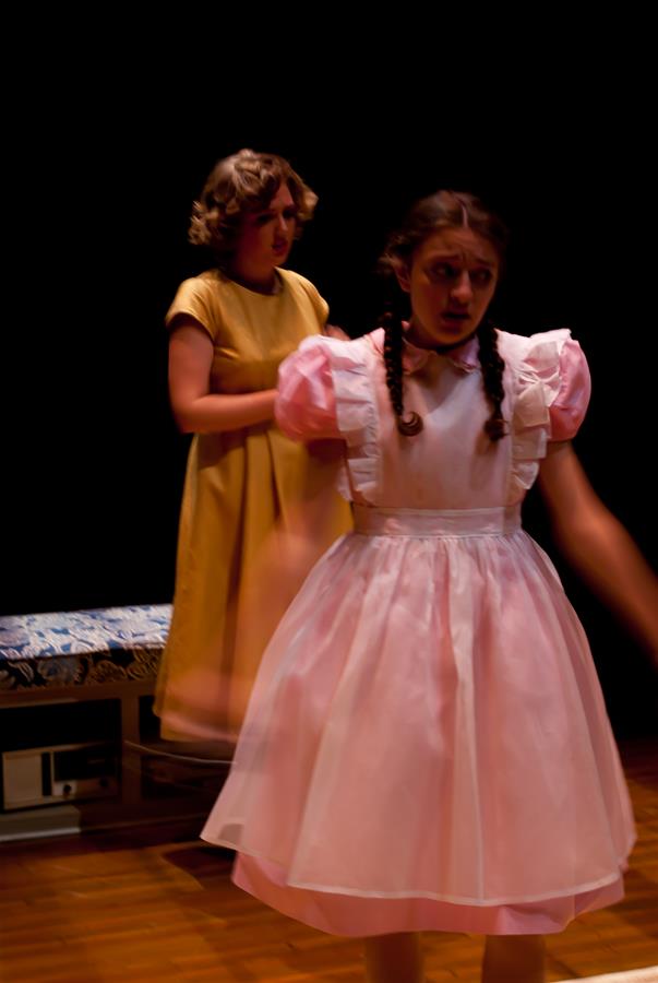 Two girls in vintage dresses are on stage. One, in a yellow dress, stands in the background, while the other, in a pink dress with puffy sleeves, appears in motion, blurred, in the foreground. The scene is set against a dark backdrop.