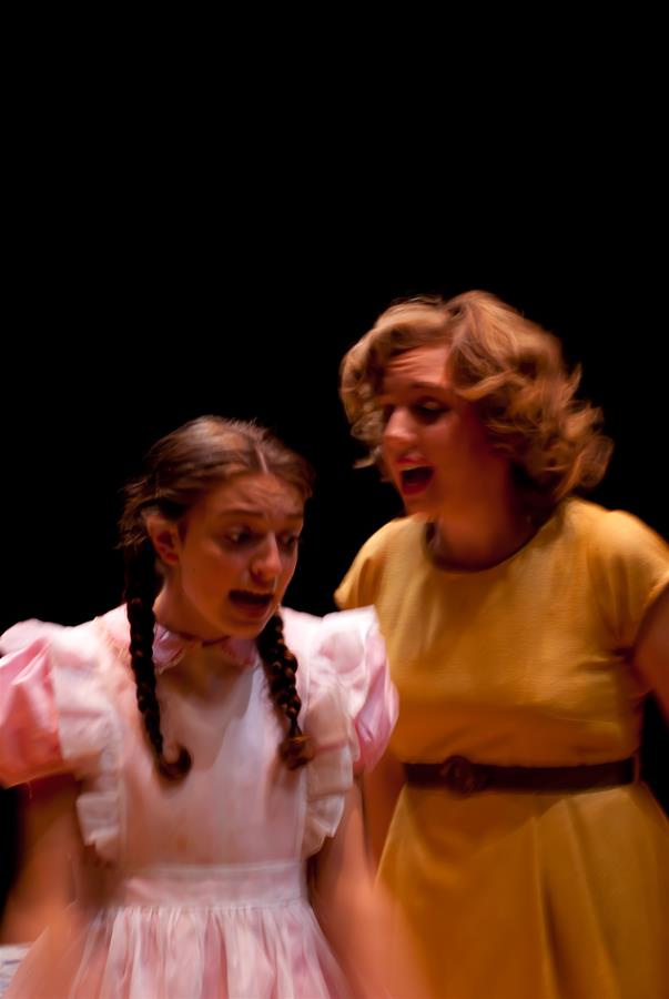 Two women are on stage in dramatic lighting. The woman on the left has braided hair and wears a pink dress, while the woman on the right has curly hair and wears a yellow dress. Both appear to be expressing emotions.