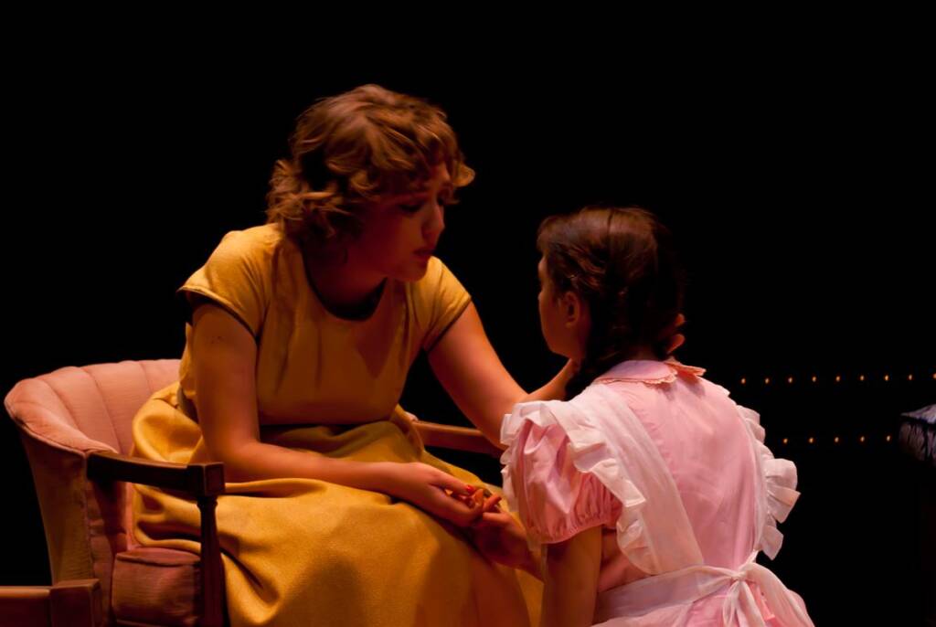 Two women sit close together on stage under dramatic lighting. The woman on the left, in a yellow dress, gently holds the hand of the woman in a pink dress with a white apron, conveying a sense of comfort and support.