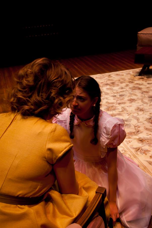 Two women indoors, one sitting in a yellow dress, facing away. The other woman, with braided hair and a pink dress, kneels on a floral rug, looking intently at the seated woman.