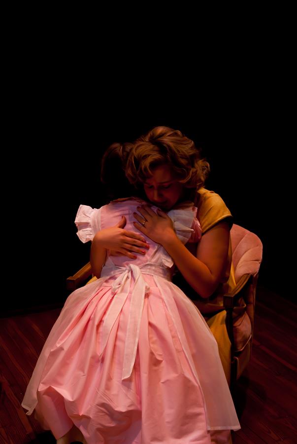 Two people sit and embrace in a warmly lit scene against a dark background. One, wearing a pink dress, hugs the other, who is in a yellow outfit. The setting suggests an intimate, comforting moment.