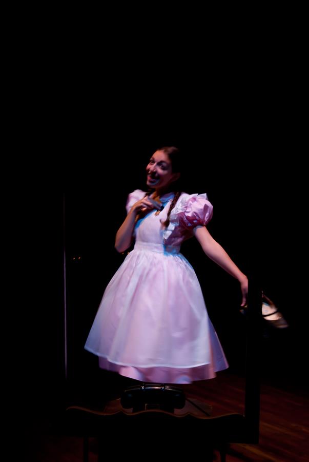 A person in a light pink dress stands on a dimly lit stage, holding a toy doll in one hand. The spotlight highlights the dress's puffed sleeves and the person's playful pose.