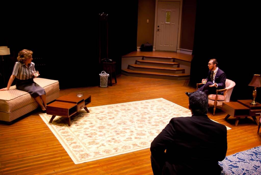 Three people are in a theater setting. A woman sits on a sofa holding a glass, and a man is seated in a chair holding a glass. Another person sits with their back to the viewer. The room has wooden floors, a rug, and a door with steps.