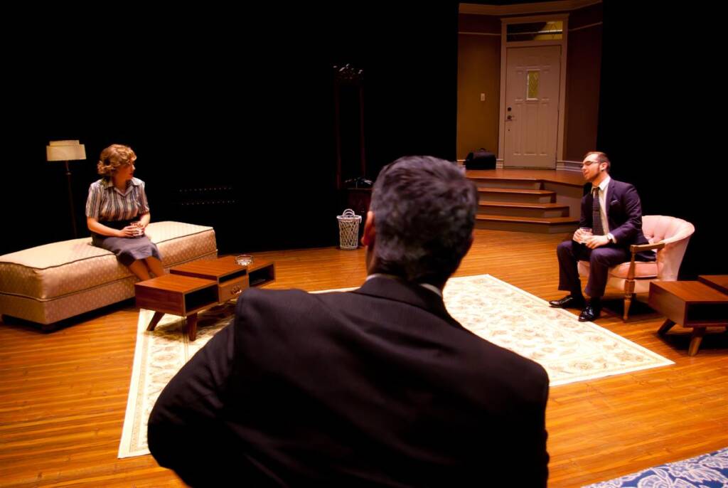 A theater scene with three actors on stage. One person is sitting on a couch, another on a chair, and a third stands with their back to the audience. The scene is set in a living room with wooden floors and dim lighting.