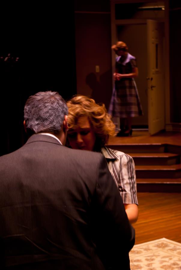 A woman with curly hair is in focus, looking down, while a man with gray hair stands closely, back to the camera. In the background, a woman in a plaid dress stands in a doorway looking on. The setting resembles a stage or a dramatic scene.