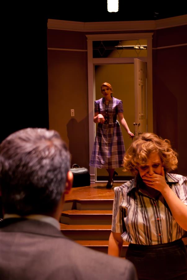 A woman in a plaid dress stands in a doorway, looking towards another woman in front who covers her mouth with her hand, appearing shocked. A man, seen from behind, faces both women. The setting is a dimly lit room with stairs.