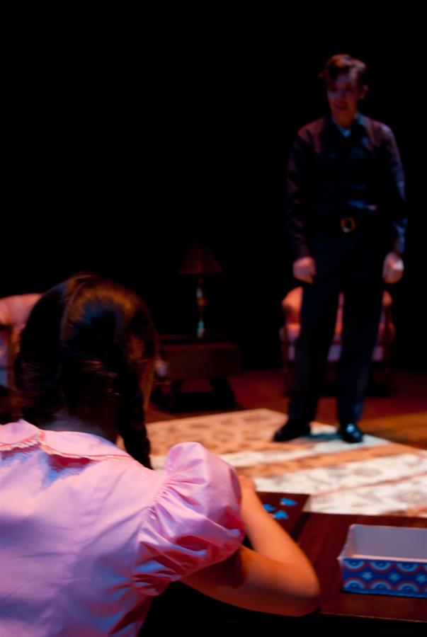 A dimly lit stage scene with a young girl in a pink dress sitting at a table, facing away, and a man in dark clothing standing in the background. A patterned rug and a lamp are visible between them.