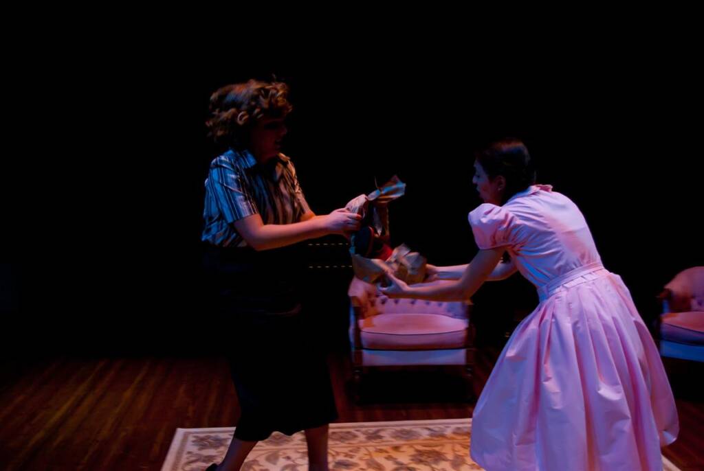 Two people are on a dimly lit stage engaged in a dramatic scene. One wears a striped shirt and dark pants, while the other is in a pink dress. They are pulling on an object in between them. There's furniture in the background.