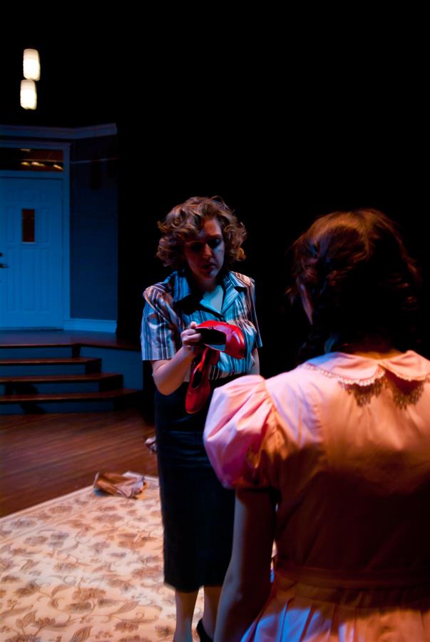 Two women are in a dimly lit room. One stands holding red shoes, facing the other, who has her back turned. A rug covers the floor, and a set of stairs leads to a door in the background. The mood appears tense.