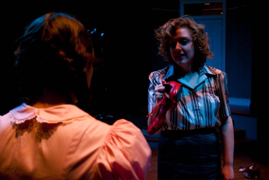 Two women in a dimly lit room. One, facing away, wears a pink blouse with lace trim. The other, facing forward, has curly hair and wears a striped blouse, holding up a red item. The atmosphere is tense.