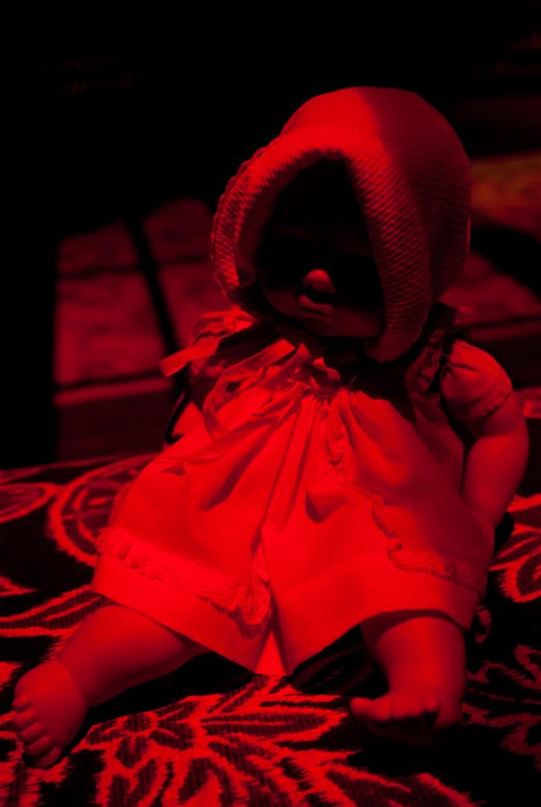 A doll wearing a bonnet and a dress is sitting under a red light, casting deep shadows. The background is dark and indistinct, adding a mysterious and eerie atmosphere to the scene.