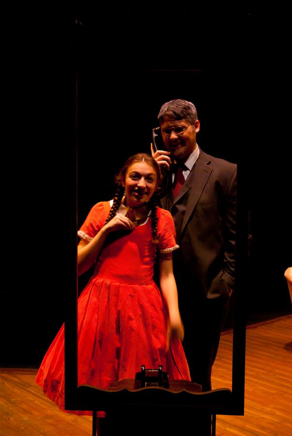 A girl in a red dress and braids stands in front of a man in a suit. They're reflected in a tall mirror on a dimly lit stage. The girl appears animated and expressive, while the man holds a phone to his ear.