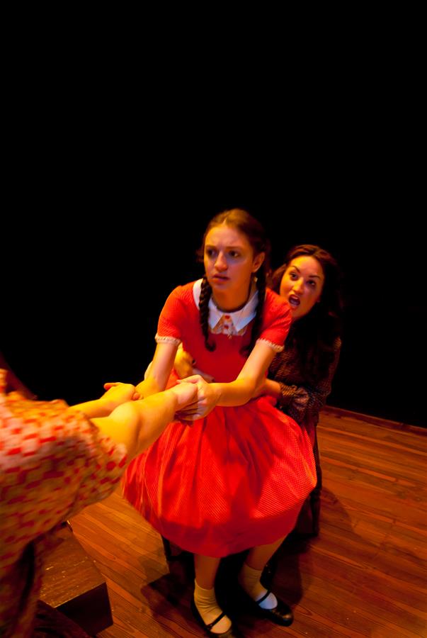 A young girl in a red dress with braided hair is being pulled by two people. She looks determined, while a woman behind her appears to be shouting. The scene is set on a dimly lit stage with a dark background.