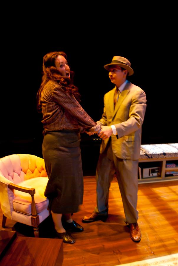 A man in a suit and hat holds hands with a woman wearing a blouse and skirt on a stage. She has long hair and appears surprised. A pink armchair is nearby. The stage is dimly lit with a warm tone.