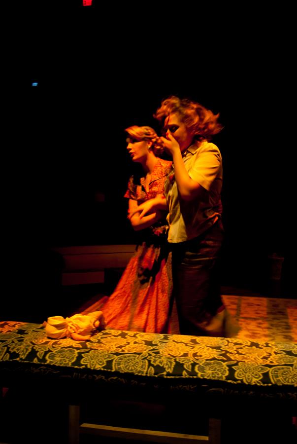 Two actors on a dimly lit stage perform an emotional scene. One wears a long, red dress and the other a casual shirt and pants. A patterned bench and small props are visible. The atmosphere is tense and dramatic.