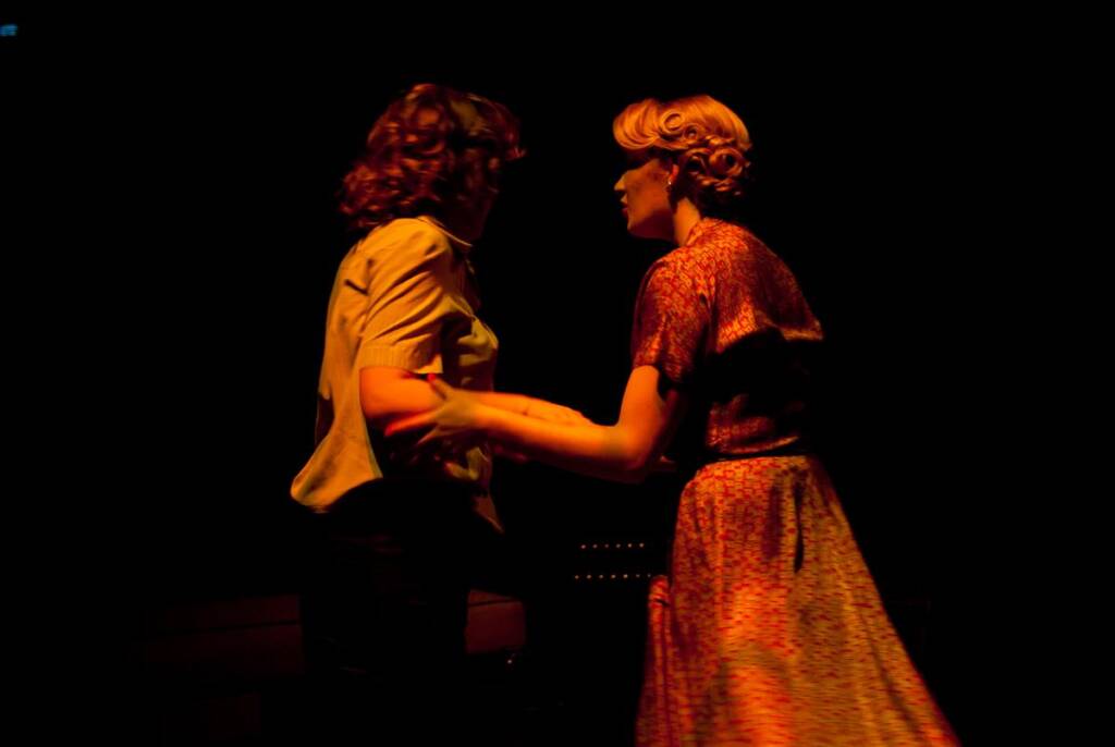 Two people stand closely together in a dimly lit setting. One wears a light shirt, and the other a patterned dress. Their expressions are not visible. The scene suggests a moment of connection or dance in a theatrical or intimate atmosphere.