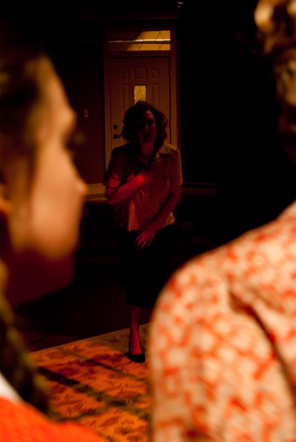 A dimly lit scene with three people. A person stands in the background, softly illuminated, wearing a shirt and dark skirt. Two other people are in the foreground, slightly out of focus, wearing patterned clothing. The mood is tense.