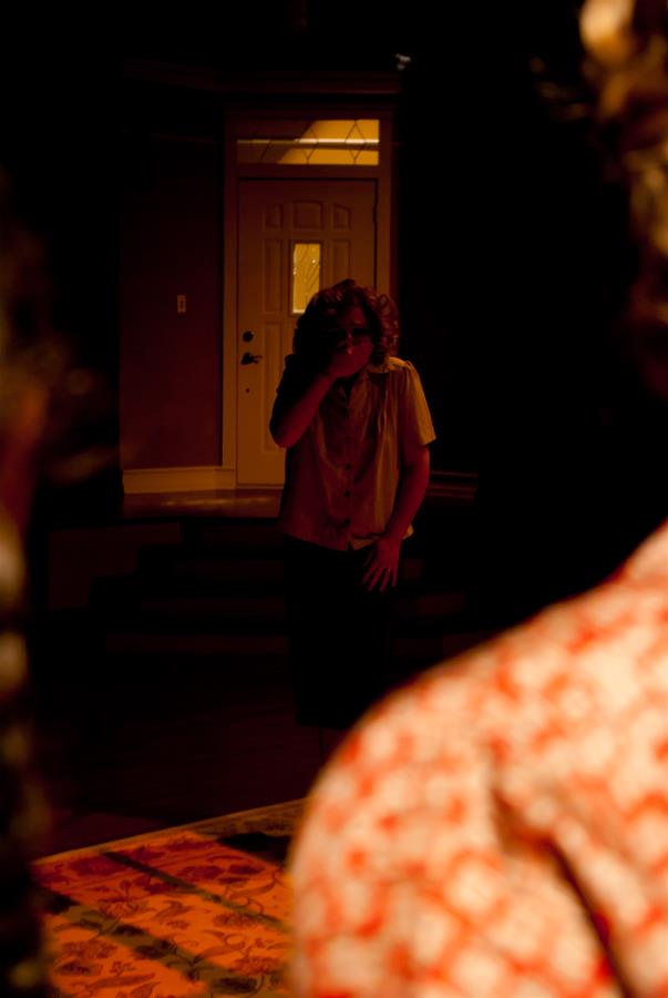 A person stands in a dimly lit room, shielding their eyes from the light. Another person, partially visible from behind, wears a patterned shirt. A door in the background is slightly ajar, casting a warm glow.