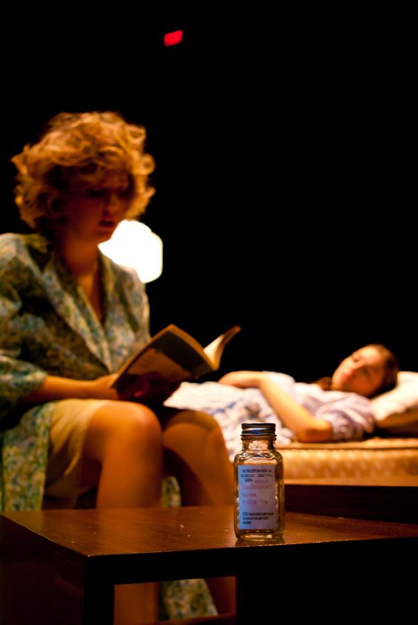 A woman with curly hair reads a book while seated next to a bed where another person lies, covered, with eyes closed. In the foreground, a small bottle is placed on a table. The scene is dimly lit.