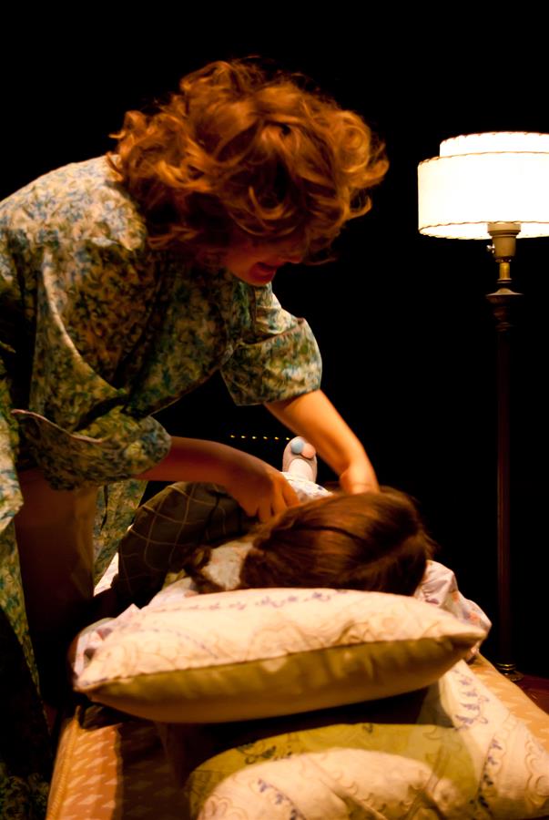 A woman with curly hair leans over a person lying on a bed, covered with pillows and a blanket. A standing lamp is lit in the background, creating a soft glow in the dimly lit room.