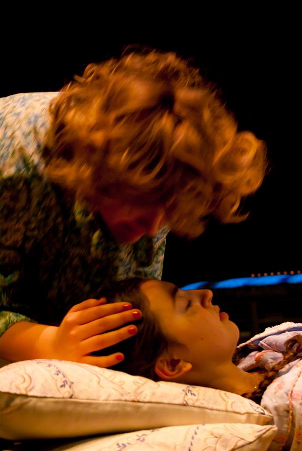 A person with curly hair leans over another person lying on a pillow, gently touching their head. The scene is dimly lit, creating a warm, intimate atmosphere.