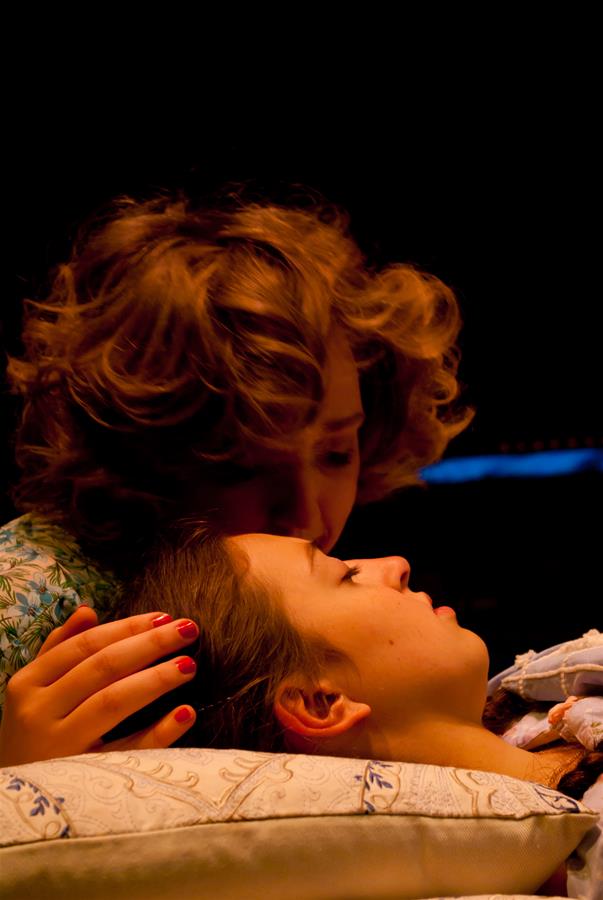 A person with curly hair gently kisses another person's forehead as they lie down with their eyes closed, both appearing peaceful and serene, under a dim, warm light.