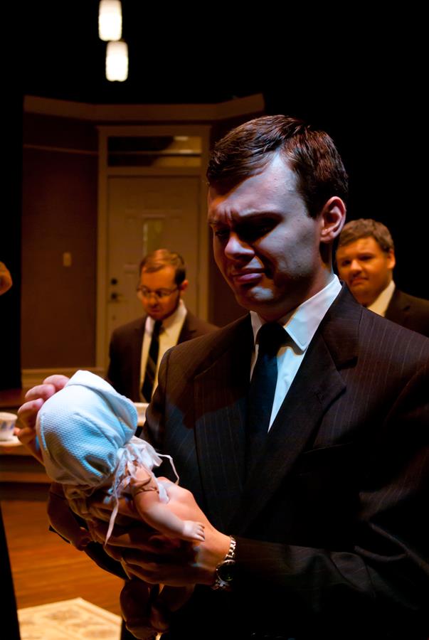 A man in a dark suit is holding a small object wrapped in white cloth, looking at it intently. Two other men in suits stand in the background, slightly out of focus. The setting appears to be indoors with warm lighting.