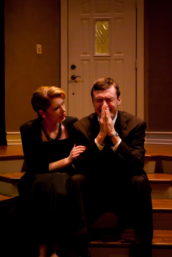 A woman and a man sit on wooden steps indoors. The woman, in a dark dress, appears concerned, with her hand on the man's shoulder. The man, in a suit, is sitting with his hands clasped over his face, looking distressed. A door is visible behind them.