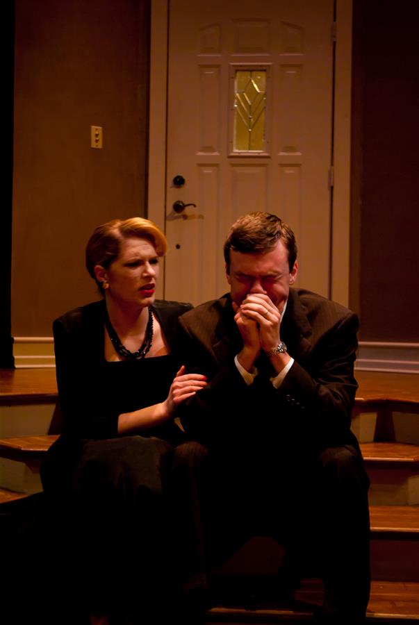 A woman and a man are sitting on steps indoors. The woman looks concerned and rests her hand on the man's arm. The man, wearing a suit, appears distressed, covering his mouth with clasped hands. A closed door is in the background.