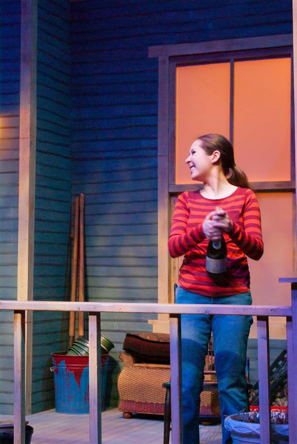 A woman in a red striped shirt and jeans joyfully opens a bottle on a wooden deck. The setting has a cozy evening vibe, with a bucket and wicker baskets in the background against a blue wall with a window.