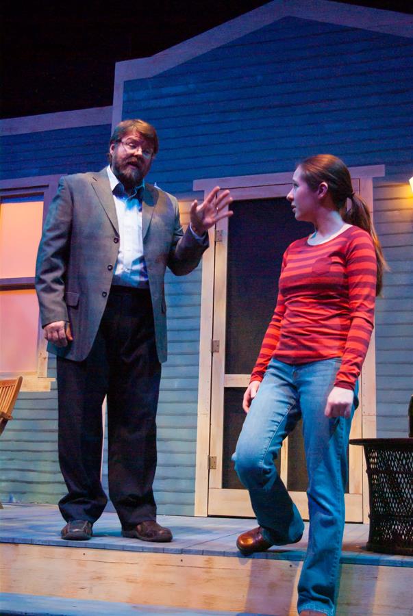 A man in a gray suit and glasses gestures while speaking to a young woman in a red striped shirt and jeans. They stand on a wooden porch in front of a house with blue siding, illuminated by warm lights.