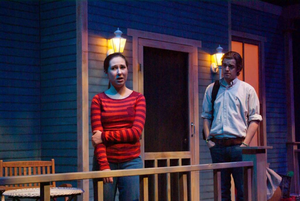 Two people stand on a dimly lit porch. The person on the left wears a red striped shirt and appears to be speaking. The person on the right, in a light shirt with rolled-up sleeves, listens with a thoughtful expression. Two lanterns illuminate the scene.