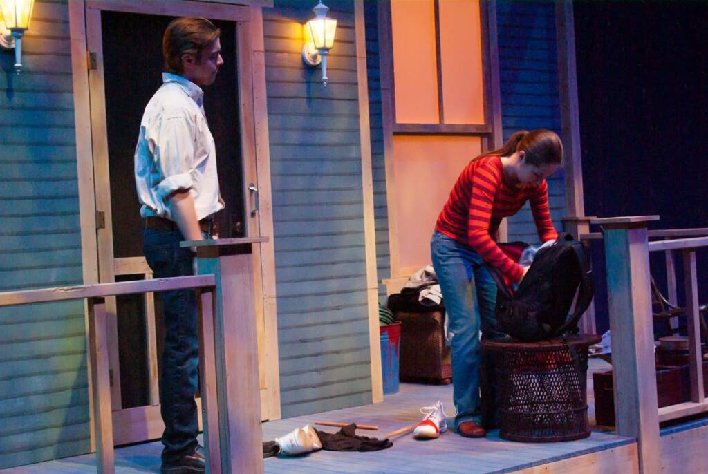 Two people stand on a porch. One, in a striped shirt, is leaning over a basket, packing a bag. The other, in a white shirt, stands nearby watching. The scene is lit by porch lights against a blue wall, suggesting evening or night.