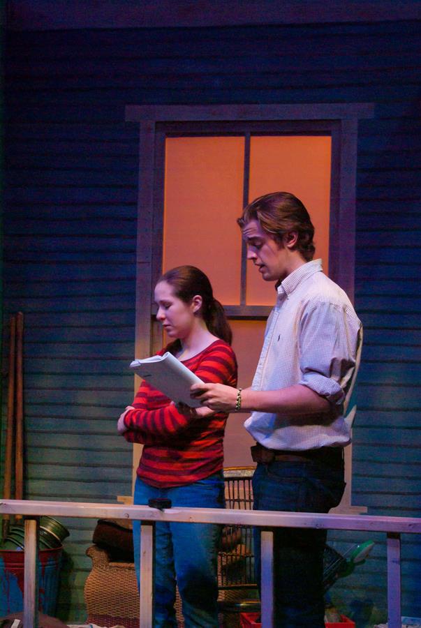 Two people stand on a porch, illuminated by warm light. The person on the left, in a red-striped sweater, listens with folded arms, while the person on the right, in a white shirt, reads from a script. A dark house facade is visible in the background.