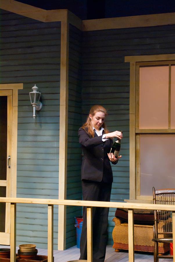 A person in a dark suit stands on a wooden porch, opening a bottle. The porch has a blue wall with a tall window and a door. There is a lantern on the wall and wicker furniture nearby.