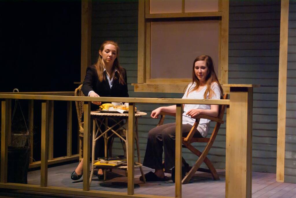 Two women sit on a porch. One wears a black jacket and is holding a notebook, while the other wears a white shirt and brown pants, seated on a folding chair. A wooden railing and table with items partially visible complete the scene.