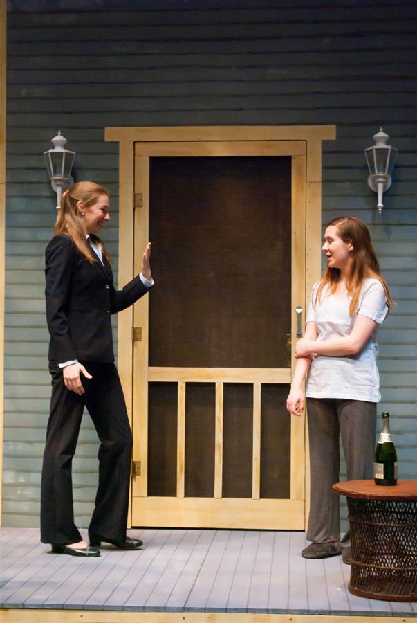 Two women stand on a porch. One is in a suit, raising her hand to greet the other, who is casually dressed in a t-shirt and pants. They face each other, with a wooden door and two wall sconces in the background. A champagne bottle is on a table nearby.