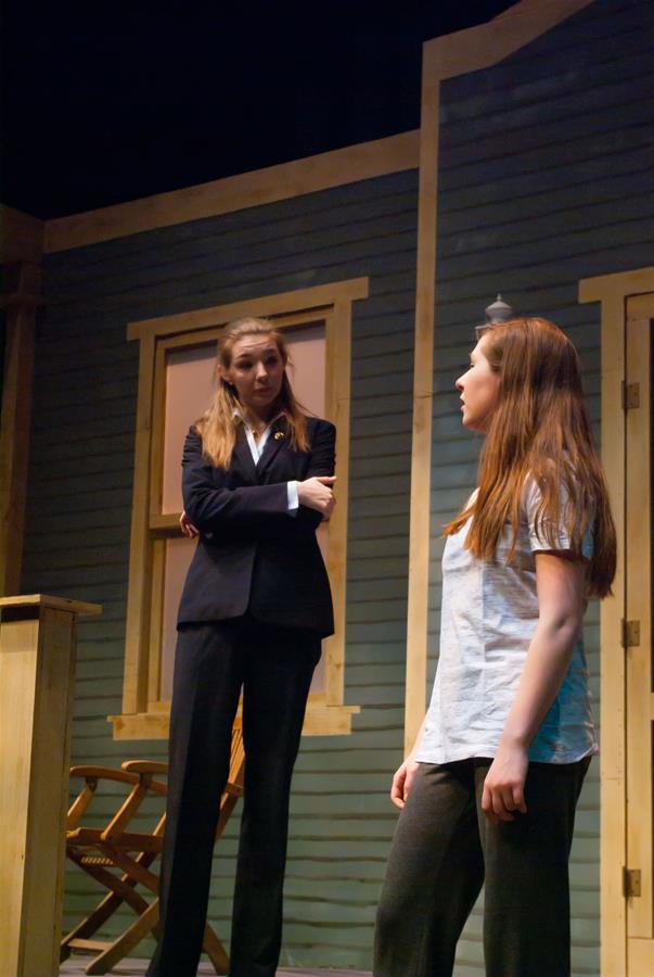 Two people are on stage performing a play. One is in a suit, looking at the other who is dressed casually in a t-shirt and pants. They are standing in front of a set designed to resemble a house with wooden walls and windows.