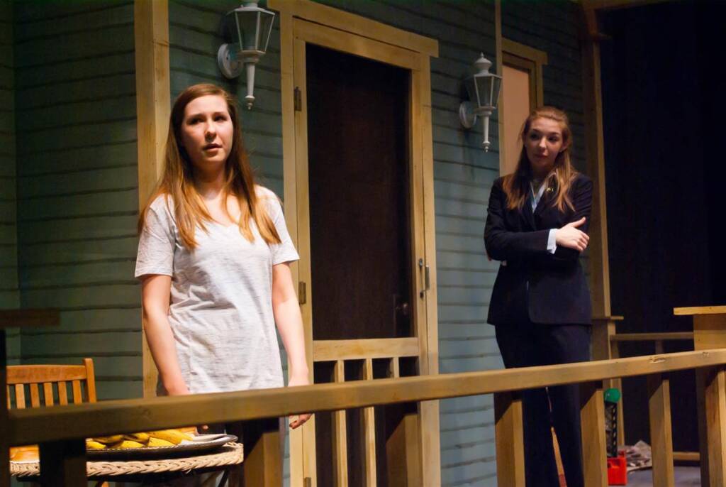 Two women stand on a wooden porch. One is wearing a gray shirt and looks thoughtful, while the other, in a black suit, stands with crossed arms. The porch has light fixtures and appears part of a house set for a play or performance.