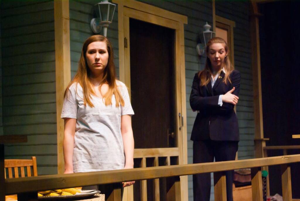 Two women stand on a porch during a stage play. One wears a white shirt and looks serious, while the other, in a dark suit, stands with folded arms. A green wall with lights, doors, and a wooden railing are in the background.