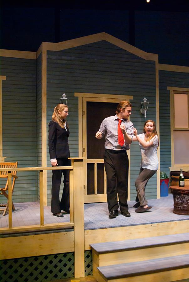 Three actors perform on a stage set resembling a house porch. A woman in a suit stands to the left, while a man in a tie grabs the wrist of a woman who appears upset. The scene includes outdoor furniture and a decorative bottle.