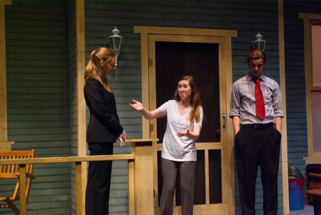 Three actors perform a scene on a theater stage. A woman gestures while speaking to another woman in a suit. A man stands nearby with his hands in his pockets. They are on the porch of a house set, with a doorway and chairs visible.