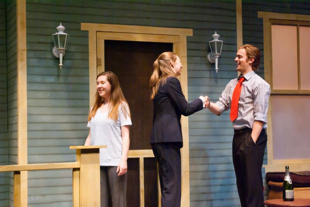 Three people on a stage in front of a house backdrop. Two are shaking hands, dressed in a suit and a shirt with a tie, respectively. The third person, in casual attire, stands beside them smiling. A bottle rests on a small table near the door.