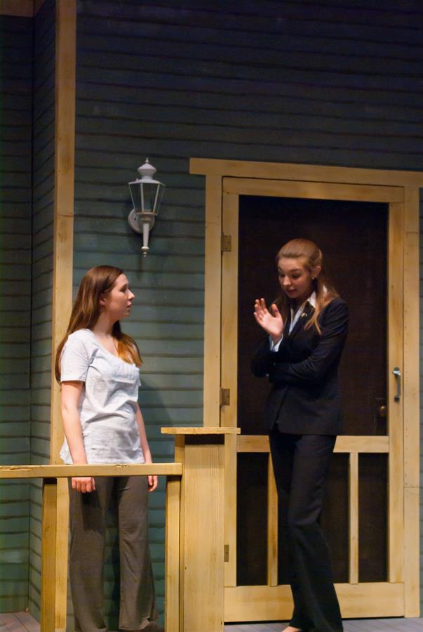 Two women stand on a porch stage set. One wears a casual outfit with a white top and gray pants. The other is in a black suit, gesturing expressively. The backdrop is a wooden door and lamp.